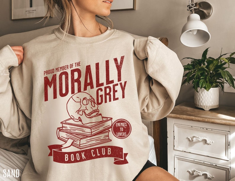 Puede incluir: Una sudadera beige con un dise&ntilde;o gr&aacute;fico rojo que dice "Proud Member of the Morally Grey Book Club". El gr&aacute;fico tambi&eacute;n incluye una calavera encima de una pila de libros y un c&iacute;rculo rojo que dice "Enemies to Lovers".