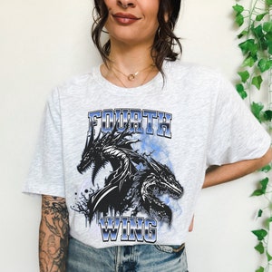 K&ouml;nnte beinhalten: Ein graues T-Shirt mit einem schwarzen und blauen Grafikdruck von drei Drachen mit dem Text "FOURTH WING" auf der Vorderseite.