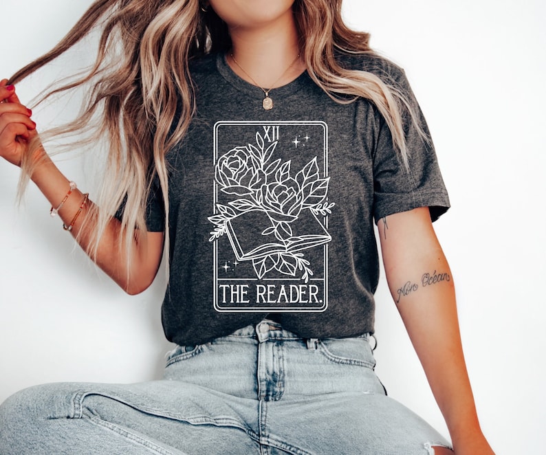 May include: Dark gray t-shirt featuring a white graphic of an open book with roses and the text "THE READER." The design is framed by a rectangular border with the Roman numeral XII at the top. The shirt is made of a soft material.