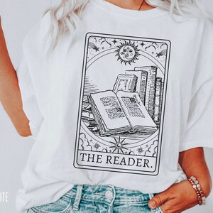 May include: White t-shirt featuring a black and white tarot card design with an open book, stacked books, and a sun and moon illustration. The text "THE READER" is printed below the image.