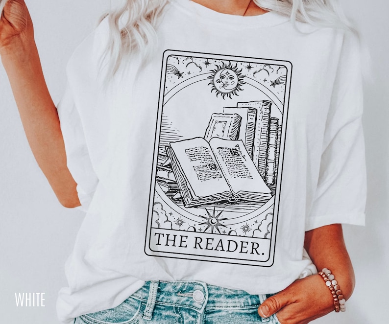 May include: White t-shirt featuring a black and white tarot card design with an open book, stacked books, and a sun and moon illustration. The text "THE READER" is printed below the image.