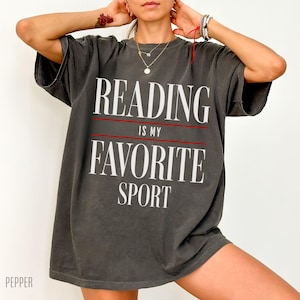 May include: A dark gray oversized t-shirt with the text "Reading is my favorite sport" printed in white on the front.