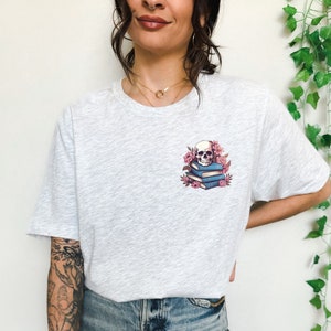 May include: Light gray t-shirt featuring a small graphic of a skull atop a stack of blue books, surrounded by pink flowers. The shirt has a crew neck and short sleeves. The word "ASH" is printed in the bottom left corner.