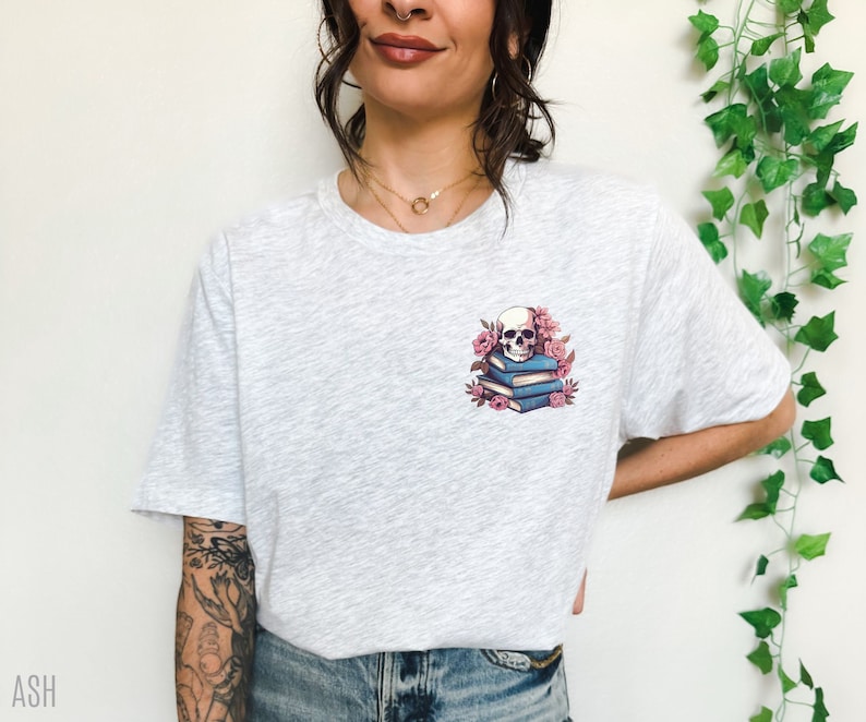 May include: Light gray t-shirt featuring a small graphic of a skull atop a stack of blue books, surrounded by pink flowers. The shirt has a crew neck and short sleeves. The word "ASH" is printed in the bottom left corner.