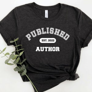 May include: A dark gray t-shirt with white text that reads "PUBLISHED EST. 2022 AUTHOR".