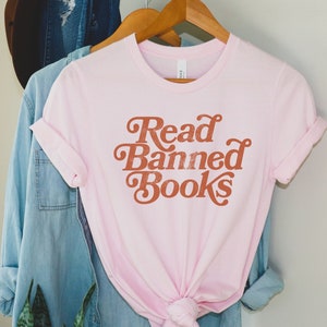 May include: Pink t-shirt with the text "Read Banned Books" in a retro font style. The text is in a burnt orange color.
