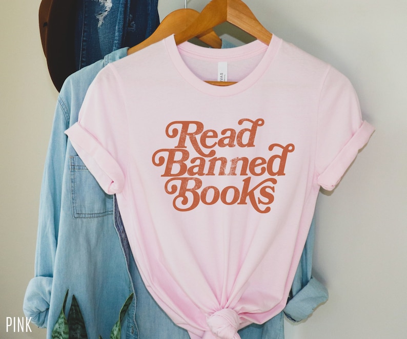 May include: Pink t-shirt with the text "Read Banned Books" in a retro font style. The text is in a burnt orange color.