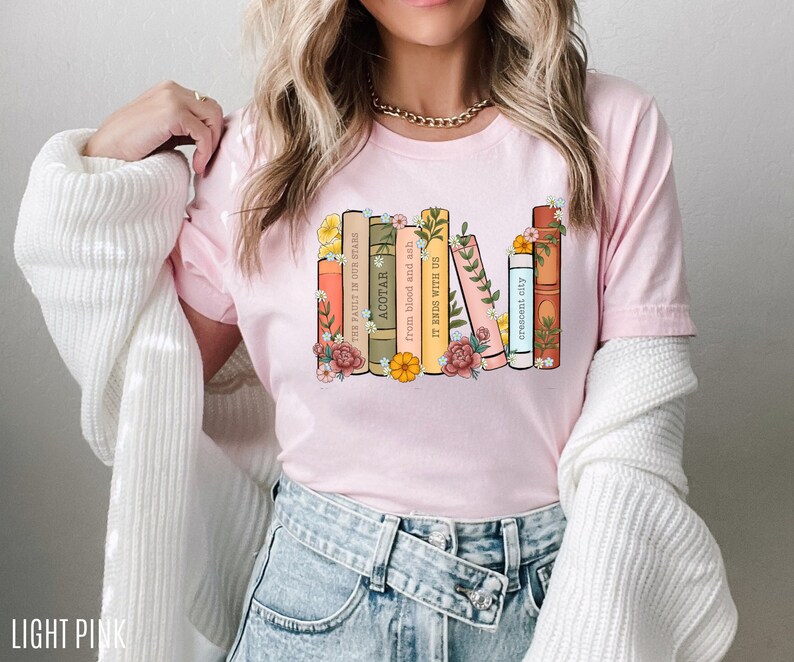 May include: A light pink t-shirt with a graphic of a stack of books with floral accents. The book titles include "The Fault in Our Stars", "ACOTAR", "From Blood and Ash", "It Ends With Us", and "Crescent City".