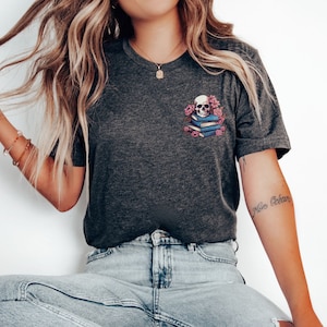 May include: Dark gray t-shirt featuring a graphic of a skull atop a stack of books, surrounded by pink flowers. The design is on the left chest area. The shirt has a crew neck and short sleeves.