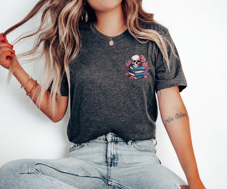 May include: Dark gray t-shirt featuring a graphic of a skull atop a stack of books, surrounded by pink flowers. The design is on the left chest area. The shirt has a crew neck and short sleeves.