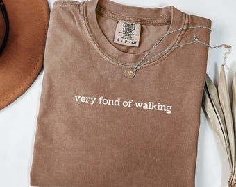 May include: Espresso-colored t-shirt with the phrase "very fond of walking" in white. A silver necklace with a pendant rests on the shirt. The shirt has a crew neck and a Comfort Colors tag.