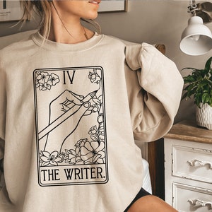 Könnte beinhalten: Ein beiger Pullover mit einer schwarz-weißen Illustration einer Tarotkarte. Die Karte ist mit "IV THE WRITER." beschriftet.