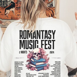 May include: White sweatshirt with the text "ROMANTASY MUSIC FEST 2024" and a skull and floral design over books. The design lists romance novel tropes. A casual, comfortable garment for a music festival.