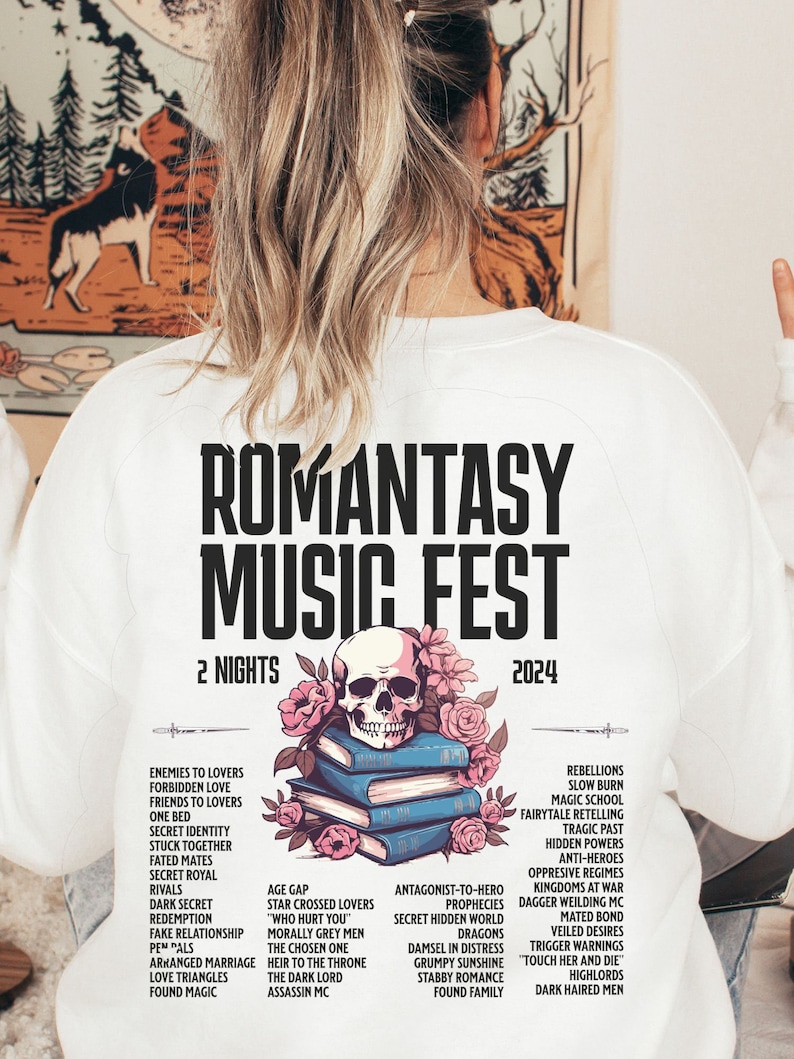 May include: White sweatshirt with the text "ROMANTASY MUSIC FEST 2024" and a skull and floral design over books. The design lists romance novel tropes. A casual, comfortable garment for a music festival.