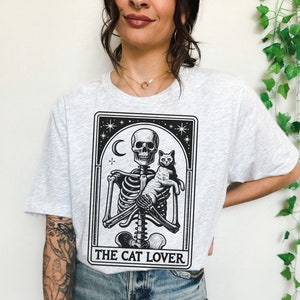 May include: Light gray t-shirt featuring a black and white graphic of a skeleton holding a cat, with the text "THE CAT LOVER" below. The design includes a crescent moon and stars. The shirt is a casual wear item.