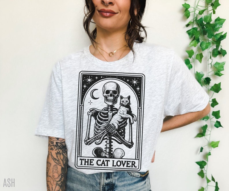 May include: Light gray t-shirt featuring a black and white graphic of a skeleton holding a cat, with the text "THE CAT LOVER" below. The design includes a crescent moon and stars. The shirt is a casual wear item.