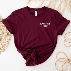 May include: A maroon t-shirt with the text "Fantasy Fest" in white with the words "Magic, Romance, Smut" in smaller white text underneath.