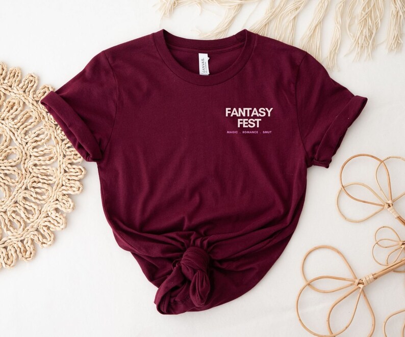 May include: A maroon t-shirt with the text "Fantasy Fest" in white with the words "Magic, Romance, Smut" in smaller white text underneath.