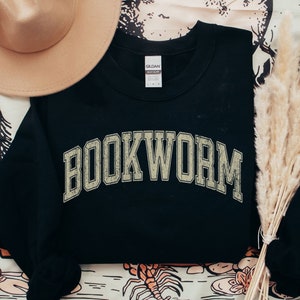 May include: Black sweatshirt with the word "BOOKWORM" in a distressed, collegiate-style font. A tan hat and dried ornamental grass are arranged near the sweatshirt. The word "BLACK" is printed at the bottom left corner.