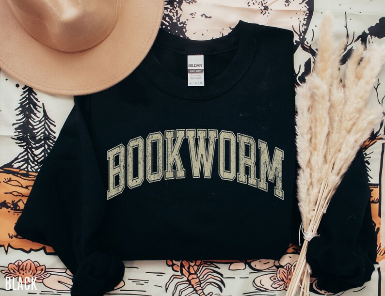 May include: Black sweatshirt with the word "BOOKWORM" in a distressed, collegiate-style font. A tan hat and dried ornamental grass are arranged near the sweatshirt. The word "BLACK" is printed at the bottom left corner.