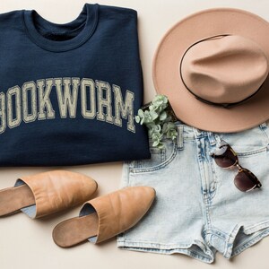May include: A navy blue sweatshirt with the word "BOOKWORM" in a distressed, arched font is displayed with a tan fedora, denim shorts, sunglasses, and tan leather slippers. The items are arranged on a neutral background, suggesting a casual, stylish outfit.