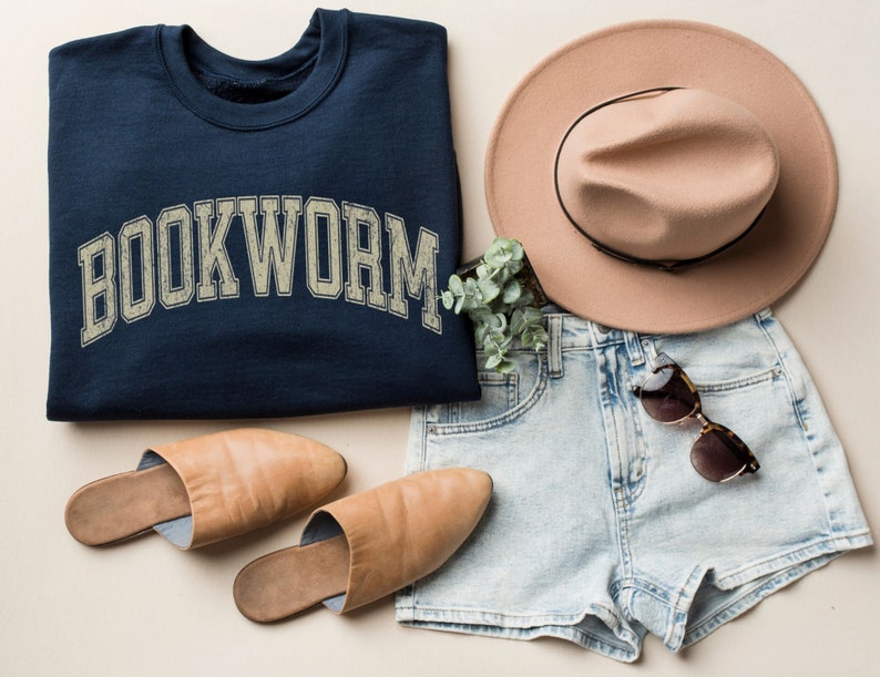 May include: A navy blue sweatshirt with the word "BOOKWORM" in a distressed, arched font is displayed with a tan fedora, denim shorts, sunglasses, and tan leather slippers. The items are arranged on a neutral background, suggesting a casual, stylish outfit.