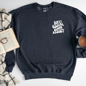 May include: Dark heather gray sweatshirt with the text "ANTI SOCIAL BOOK ADDICT" in white. The sweatshirt is next to an open book with glasses and a mug of coffee. A cream and black plaid blanket is also in the image.
