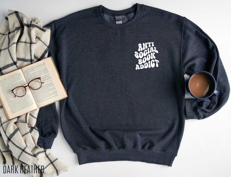 May include: Dark heather gray sweatshirt with the text "ANTI SOCIAL BOOK ADDICT" in white. The sweatshirt is next to an open book with glasses and a mug of coffee. A cream and black plaid blanket is also in the image.