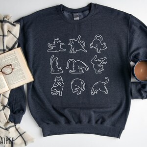 May include: Dark heather grey sweatshirt with a white outline design of cats in yoga positions. The text "DARK HEATHER" is at the bottom left. A comfortable, casual top for cat enthusiasts.