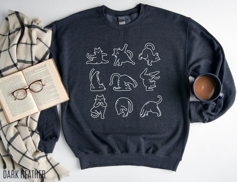 May include: Dark heather grey sweatshirt with a white outline design of cats in yoga positions. The text "DARK HEATHER" is at the bottom left. A comfortable, casual top for cat enthusiasts.