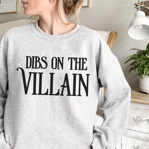 K&ouml;nnte beinhalten: Hellgraues Sweatshirt mit dem schwarzen Text "DIBS ON THE VILLAIN". Das Sweatshirt ist ein l&auml;ssiger Stil, geeignet f&uuml;r den Alltag. Das Wort "ASH" ist unten links aufgedruckt.