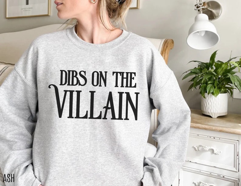 K&ouml;nnte beinhalten: Hellgraues Sweatshirt mit dem schwarzen Text "DIBS ON THE VILLAIN". Das Sweatshirt ist ein l&auml;ssiger Stil, geeignet f&uuml;r den Alltag. Das Wort "ASH" ist unten links aufgedruckt.