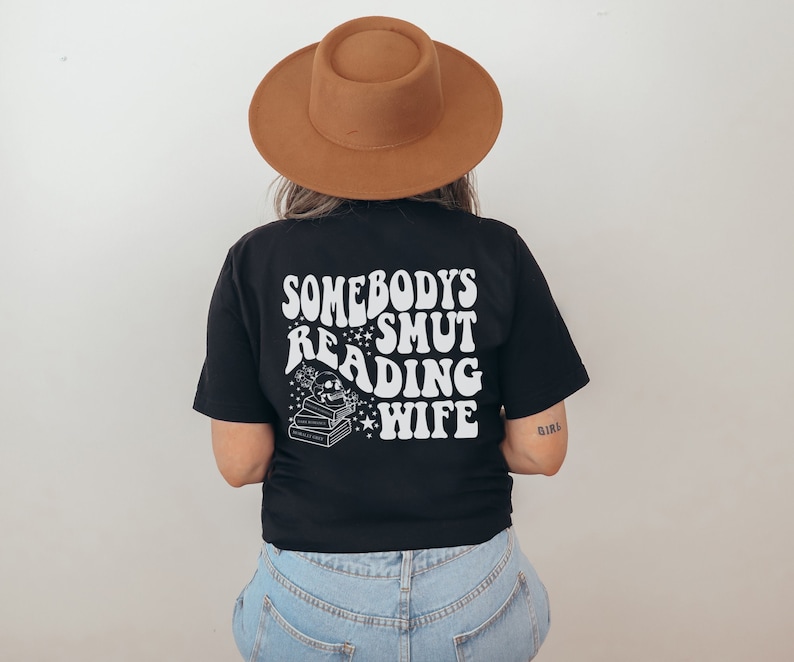 Puede incluir: Camiseta negra con el texto blanco "SOMEBODY'S SMUT READING WIFE", junto con un gr&aacute;fico de calavera, libros y estrellas. La persona lleva un sombrero marr&oacute;n. La camiseta es de estilo casual.