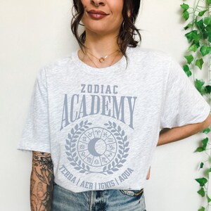 May include: Light grey t-shirt with the text "ZODIAC ACADEMY" in a stylized font, surrounded by a wreath and zodiac symbols. The shirt also features the words "TERRA AER IGNIS AQUA" below the wreath. The shirt is made of a soft material.