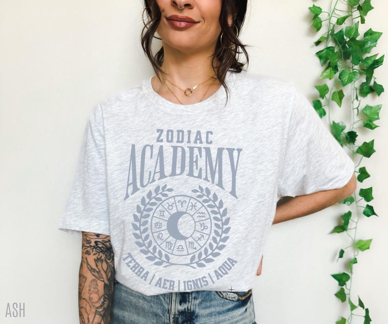 May include: Light grey t-shirt with the text "ZODIAC ACADEMY" in a stylized font, surrounded by a wreath and zodiac symbols. The shirt also features the words "TERRA AER IGNIS AQUA" below the wreath. The shirt is made of a soft material.