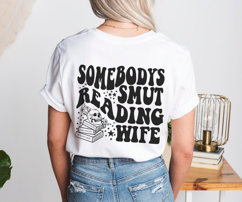 Puede incluir: Camiseta blanca con un dise&ntilde;o gr&aacute;fico negro en la espalda. El dise&ntilde;o incluye el texto "SOMEBODY'S SMUT READING WIFE" en una fuente retro, junto con una calavera, flores, estrellas y una pila de libros.