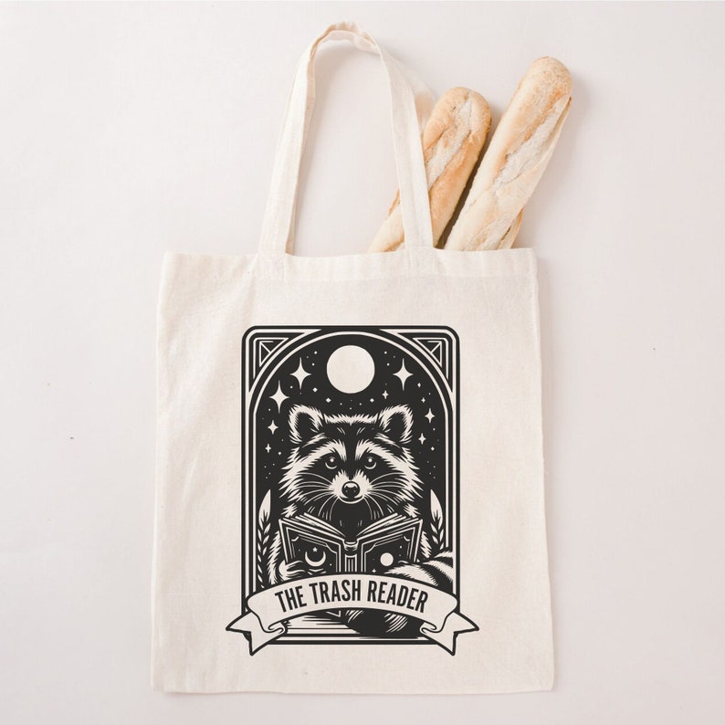 May include: Cream-colored tote bag with a black and white graphic of a raccoon reading a book, with the text "THE TRASH READER" on a banner. Two baguettes are inside the bag. The bag has two handles.