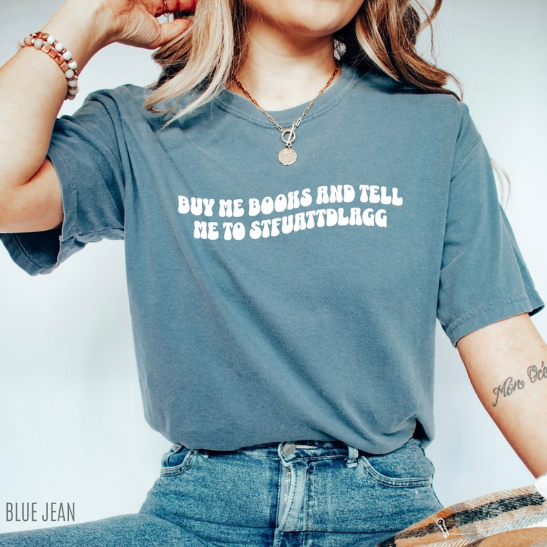 May include: A blue jean colored t-shirt with the text "BUY ME BOOKS AND TELL ME TO STFUATTDLAGG" in white. The shirt is a crew neck style and is worn with blue jeans. A gold necklace and bracelet are also visible.