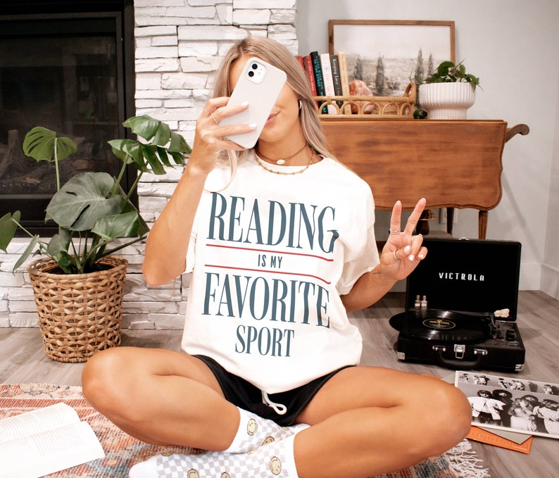 Może przedstawiać: Biała koszulka z nadrukiem "Reading is my favorite sport" w kolorze niebieskim i czerwonym. Osoba w koszulce siedzi na podłodze z książką i gramofonem.