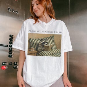 Woman wearing white oversized t shirt with probably reading trash text and raccoon reading book graphic, photographed in elevator setting