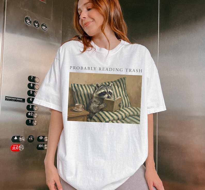 Woman wearing white oversized t shirt with probably reading trash text and raccoon reading book graphic, photographed in elevator setting