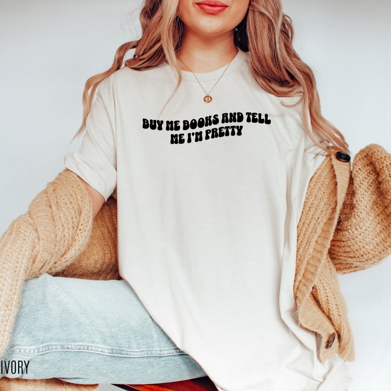 May include: A white t-shirt with the text "Buy me books and tell me I'm pretty" in black, retro-style lettering.