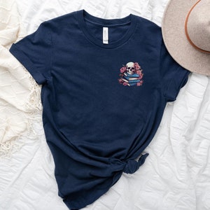 May include: Navy blue t-shirt featuring a small graphic of a skull atop a stack of books, surrounded by pink flowers. The shirt has short sleeves and a crew neck. A beige hat is visible in the upper right corner. The word "NAVY" is printed at the bottom left.