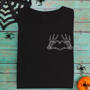 May include: Black t-shirt featuring a white skeleton hand design forming a heart shape on the chest. The shirt is folded and placed on a teal and brown wooden surface, with Halloween decorations like spiders and pumpkins.