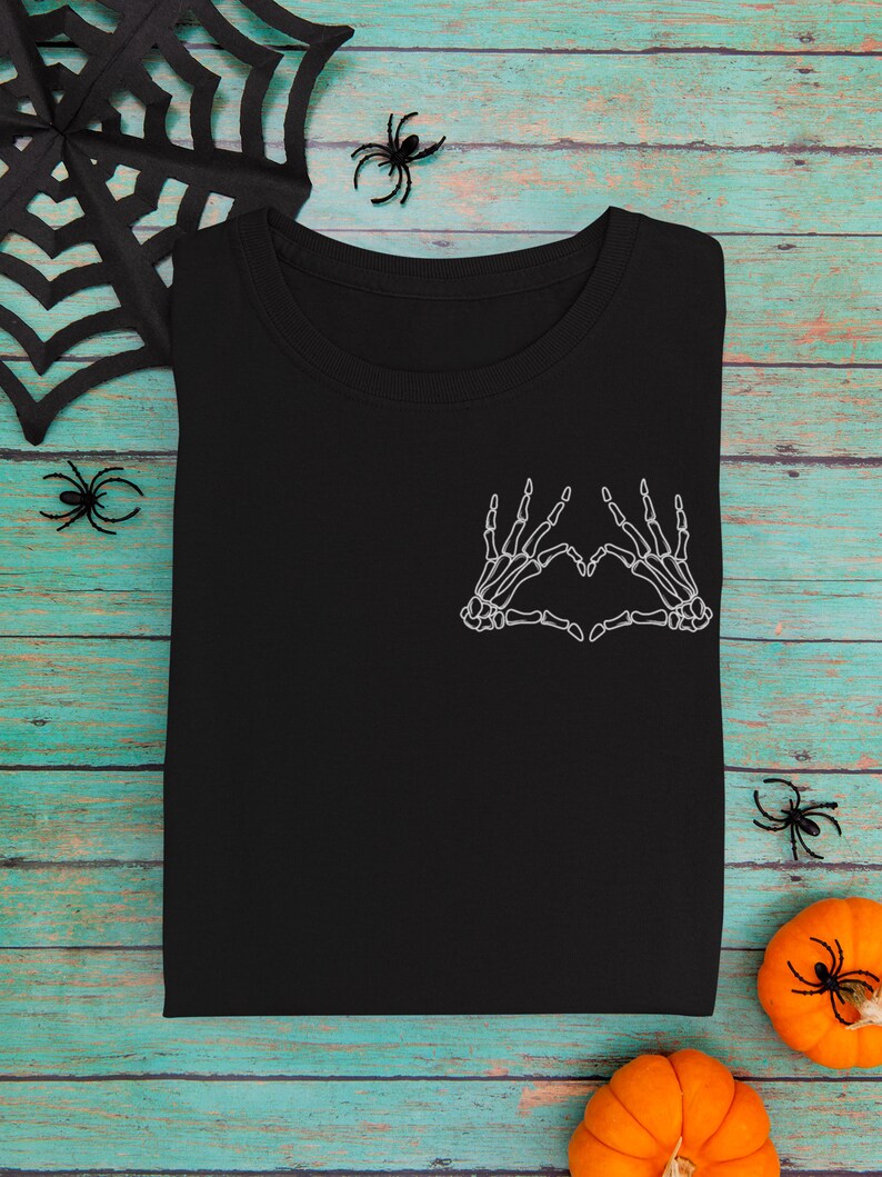 May include: Black t-shirt featuring a white skeleton hand design forming a heart shape on the chest. The shirt is folded and placed on a teal and brown wooden surface, with Halloween decorations like spiders and pumpkins.