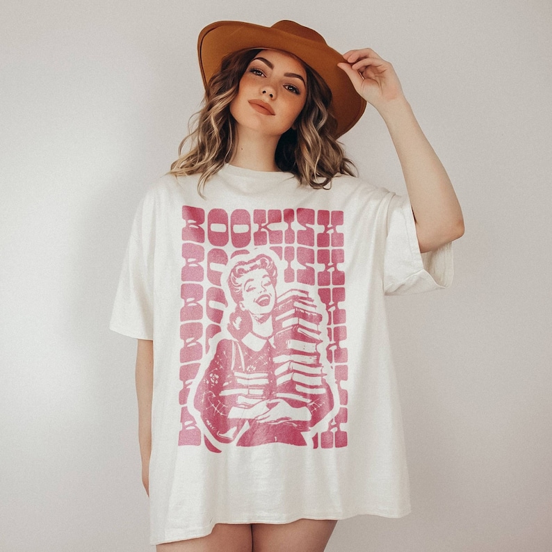 May include: A white oversized t-shirt with a pink graphic of a woman holding a stack of books. The graphic has the word "BOOKISH" printed in pink letters.