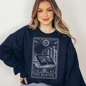 Puede incluir: Una sudadera azul marino con un gr&aacute;fico blanco de una carta de tarot que representa un libro con el texto "THE READER."