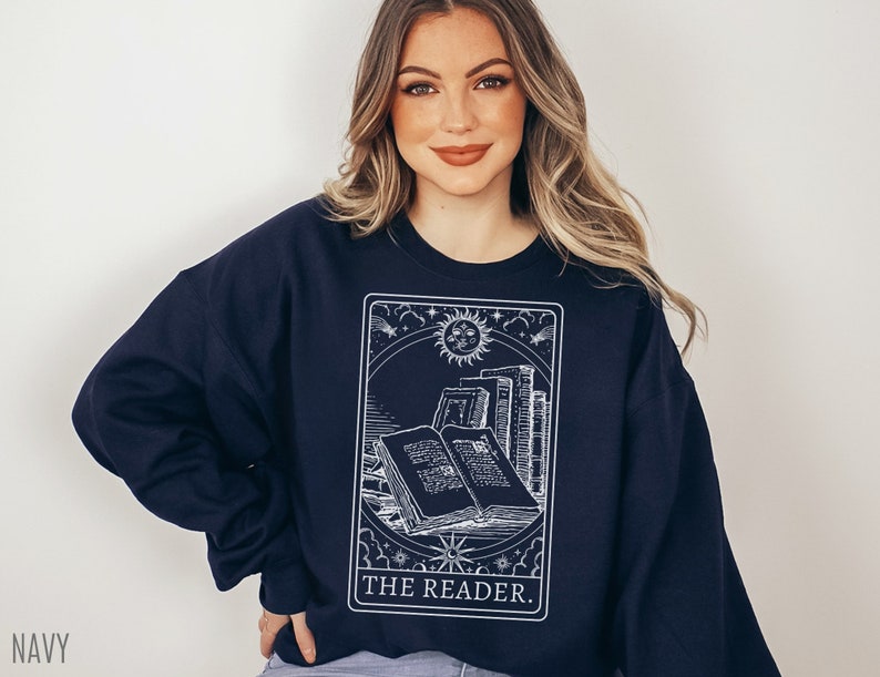 Puede incluir: Una sudadera azul marino con un gr&aacute;fico blanco de una carta de tarot que representa un libro con el texto "THE READER."