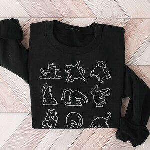 May include: Black sweatshirt with white line drawings of cats in yoga positions. The word "BLACK" is printed in white at the bottom left. The top has long sleeves and a crew neck.