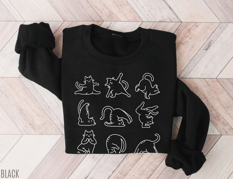 May include: Black sweatshirt with white line drawings of cats in yoga positions. The word "BLACK" is printed in white at the bottom left. The top has long sleeves and a crew neck.